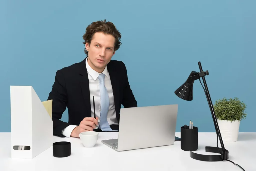 Homem em traje formal sentado à mesa com laptop, segurando uma caneta, em um ambiente corporativo moderno e organizado