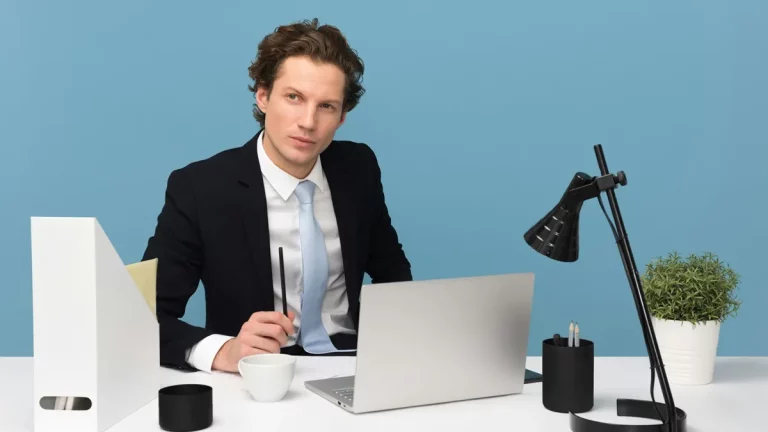 Homem em traje formal sentado à mesa com laptop, segurando uma caneta, em um ambiente corporativo moderno e organizado