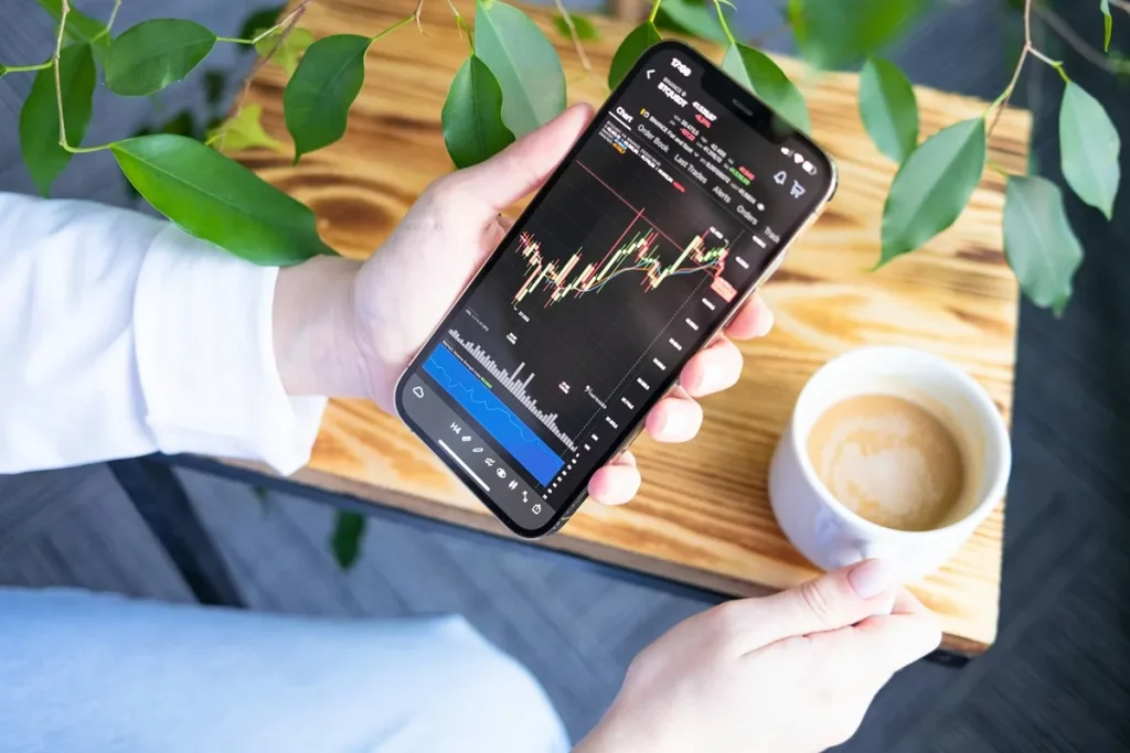 Pessoa segurando um smartphone exibindo gráfico de mercado financeiro, com uma xícara de café sobre uma mesa de madeira e folhas verdes ao fundo.