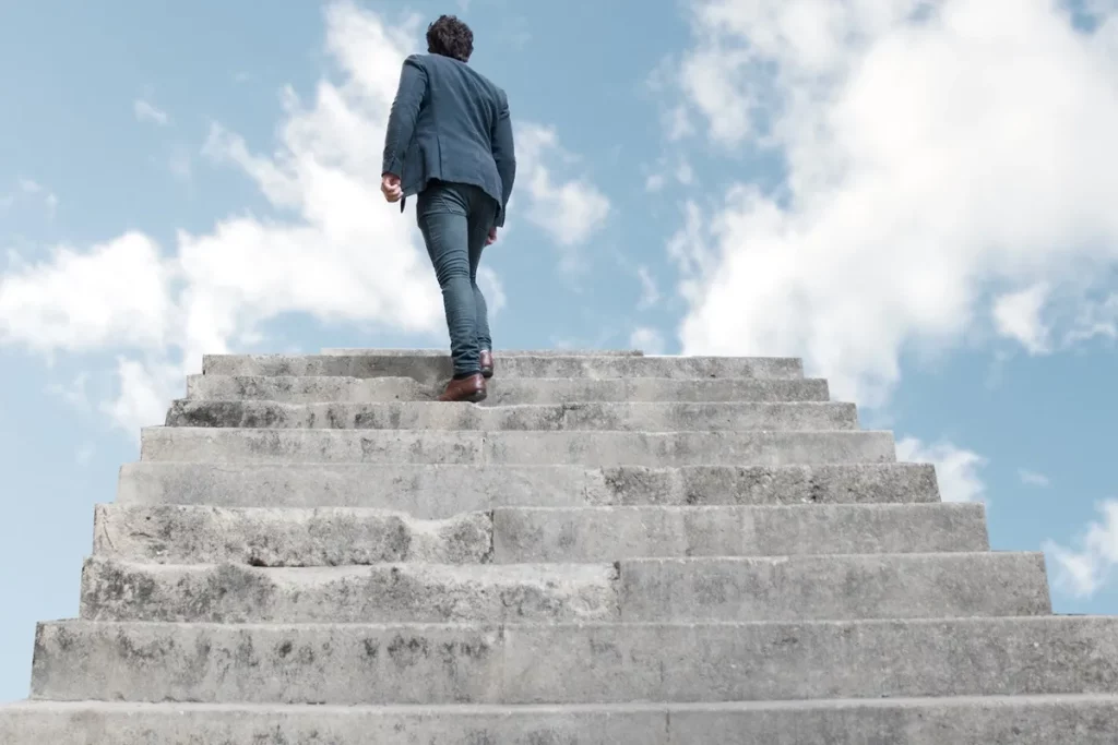 Pessoa subindo escadaria com fundo de céu azul, representando o progresso em direção às metas.