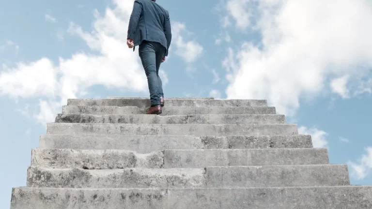 Pessoa subindo escadaria com fundo de céu azul, representando o progresso em direção às metas.