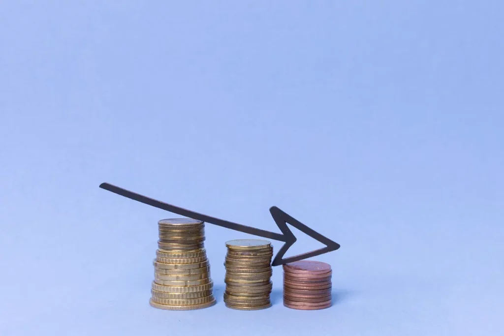 Três pilhas de moedas em diferentes alturas com uma seta preta apontando para baixo, representando declínio financeiro e perda de valor de ativos.