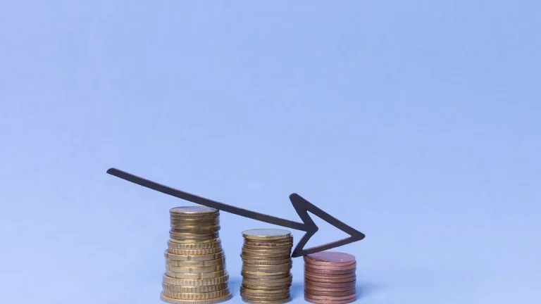 Três pilhas de moedas em diferentes alturas com uma seta preta apontando para baixo, representando declínio financeiro e perda de valor de ativos.