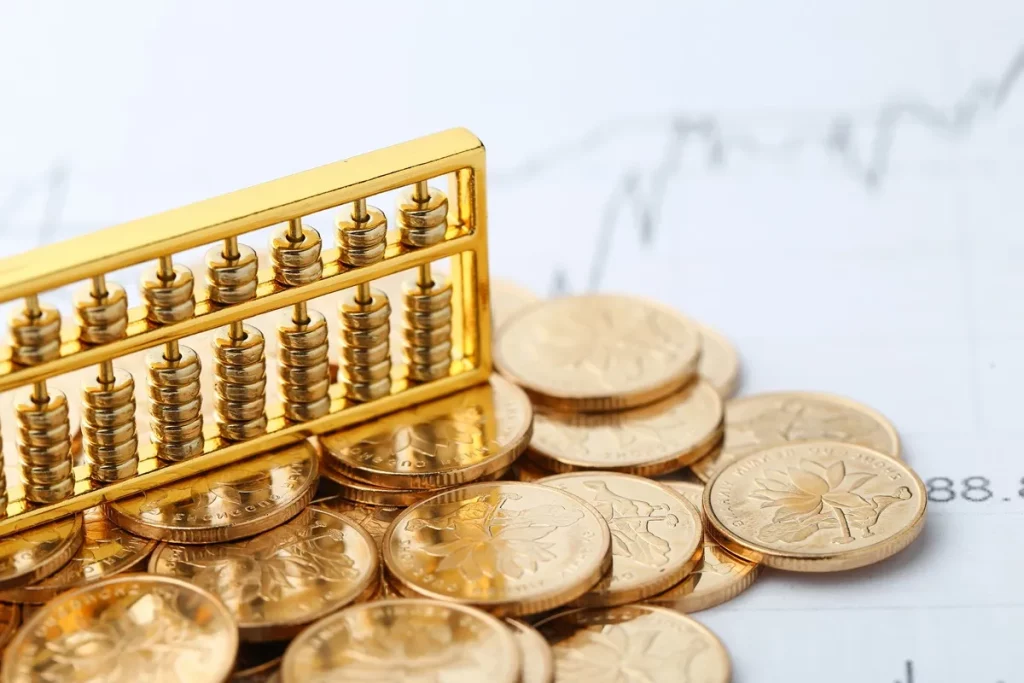 Conjunto de moedas douradas empilhadas ao lado de um ábaco dourado com contas metálicas sobre uma superfície com gráficos financeiros ao fundo.