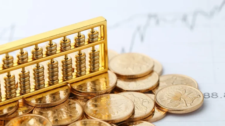 Conjunto de moedas douradas empilhadas ao lado de um ábaco dourado com contas metálicas sobre uma superfície com gráficos financeiros ao fundo.
