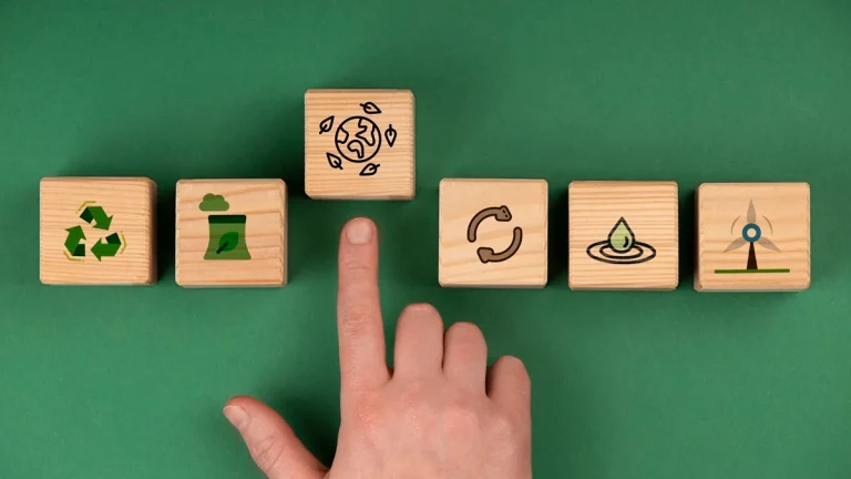 Mão apontando para um bloco de madeira com ícone de sustentabilidade ao lado de outros blocos com símbolos de reciclagem, energia limpa, e reaproveitamento em fundo verde