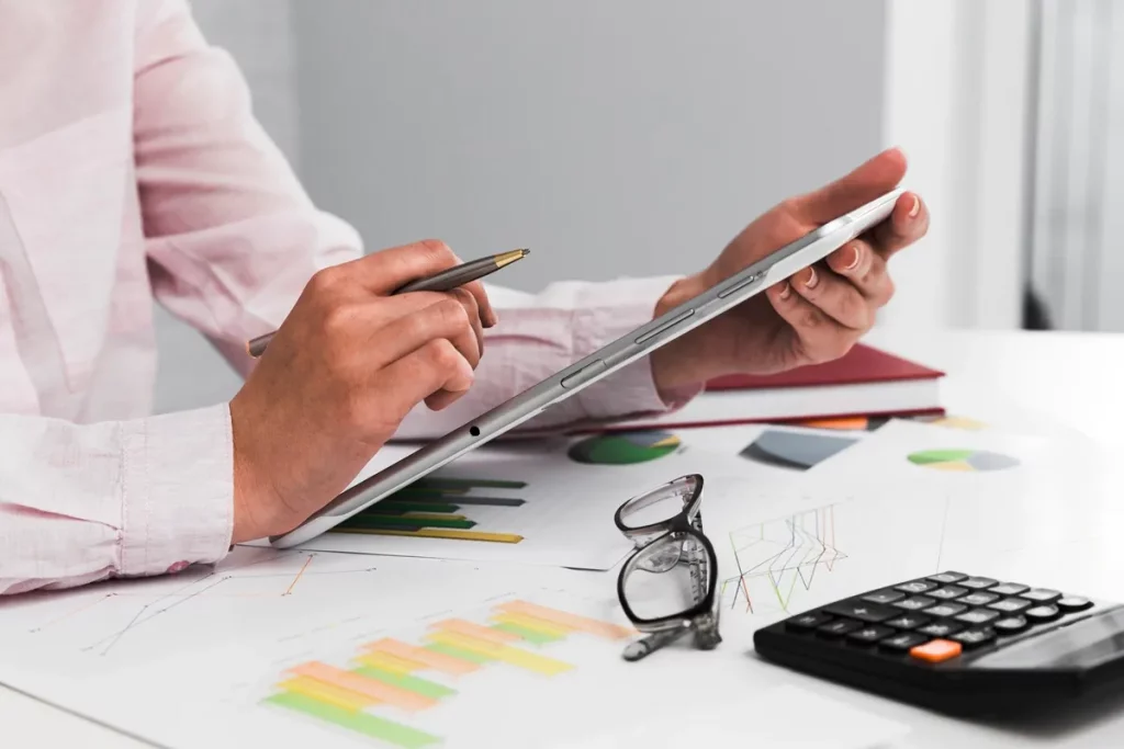 Pessoa segurando um tablet e uma caneta sobre uma mesa com gráficos de análise financeira, calculadora e óculos, representando gestão de finanças e planejamento estratégico."