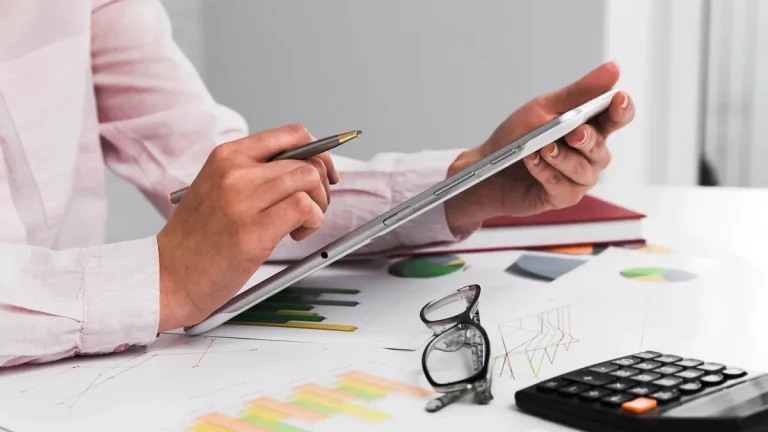 Pessoa segurando um tablet e uma caneta sobre uma mesa com gráficos de análise financeira, calculadora e óculos, representando gestão de finanças e planejamento estratégico."