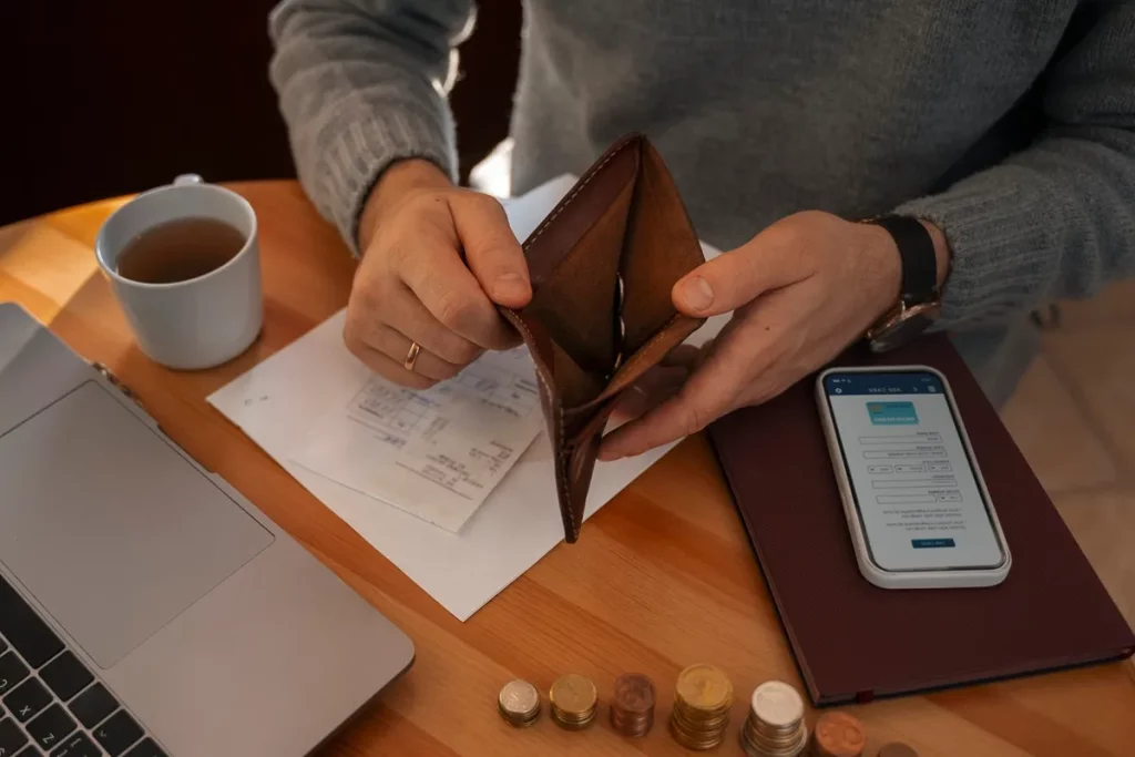 Pessoa analisando documentos financeiros enquanto segura uma carteira vazia, com moedas, um celular e um laptop sobre a mesa.