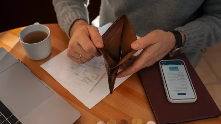 Pessoa analisando documentos financeiros enquanto segura uma carteira vazia, com moedas, um celular e um laptop sobre a mesa.