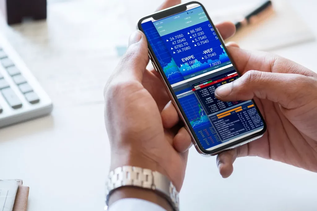 Pessoa utilizando um smartphone com uma tela exibindo gráficos e cotações de ações em tempo real, com interface de home broker e fundo com elementos de escritório, como teclado e caderno.