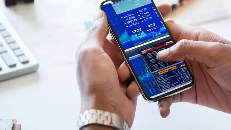 Pessoa utilizando um smartphone com uma tela exibindo gráficos e cotações de ações em tempo real, com interface de home broker e fundo com elementos de escritório, como teclado e caderno.