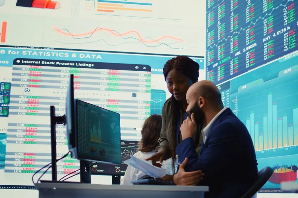 Equipe analisando gráficos de investimentos em tela de computador, cercados por grandes painéis com cotações e estatísticas de mercado financeiro