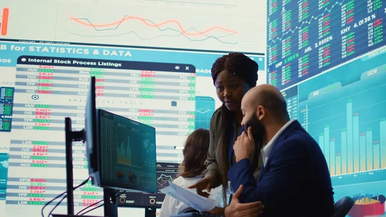 Equipe analisando gráficos de investimentos em tela de computador, cercados por grandes painéis com cotações e estatísticas de mercado financeiro
