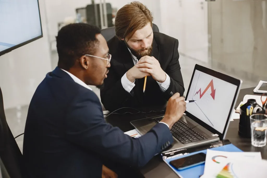 Dois homens em um ambiente corporativo analisam um gráfico em um laptop, discutindo tendências de mercado e estratégias.