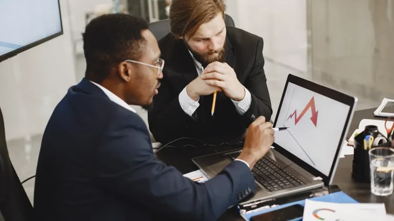 Dois homens em um ambiente corporativo analisam um gráfico em um laptop, discutindo tendências de mercado e estratégias.