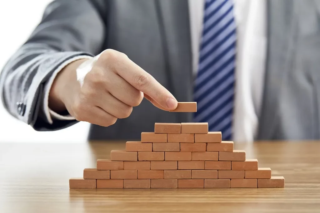 Mão de um homem de terno colocando a peça final no topo de uma pirâmide feita de pequenos blocos de madeira, representando estratégia e crescimento financeiro.