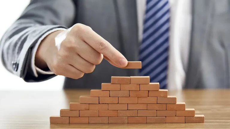 Mão de um homem de terno colocando a peça final no topo de uma pirâmide feita de pequenos blocos de madeira, representando estratégia e crescimento financeiro.