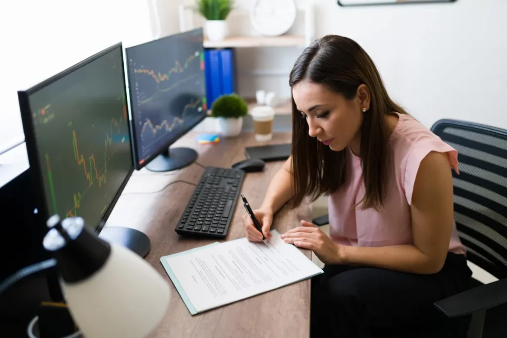 Mulher analisando gráficos financeiros em dois monitores enquanto preenche documentos, representando gestão de investimentos e planejamento financeiro.