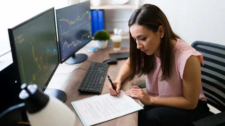 Mulher analisando gráficos financeiros em dois monitores enquanto preenche documentos, representando gestão de investimentos e planejamento financeiro.