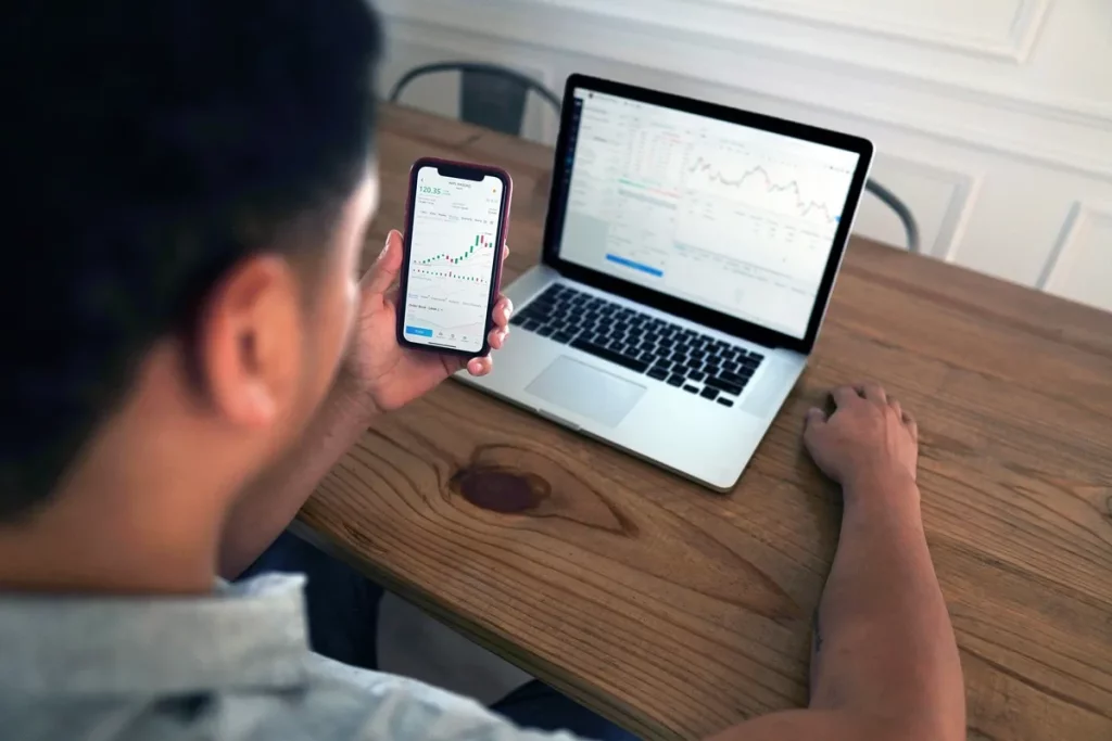 Pessoa observando gráficos de ações em celular, com notebook exibindo dados financeiros sobre a mesa