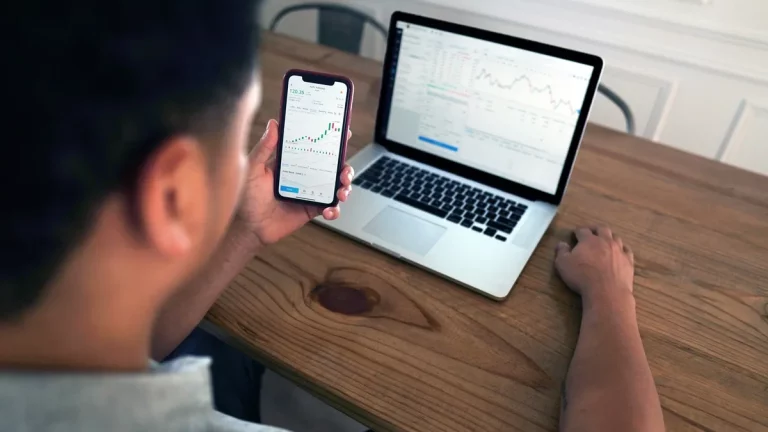 Pessoa observando gráficos de ações em celular, com notebook exibindo dados financeiros sobre a mesa