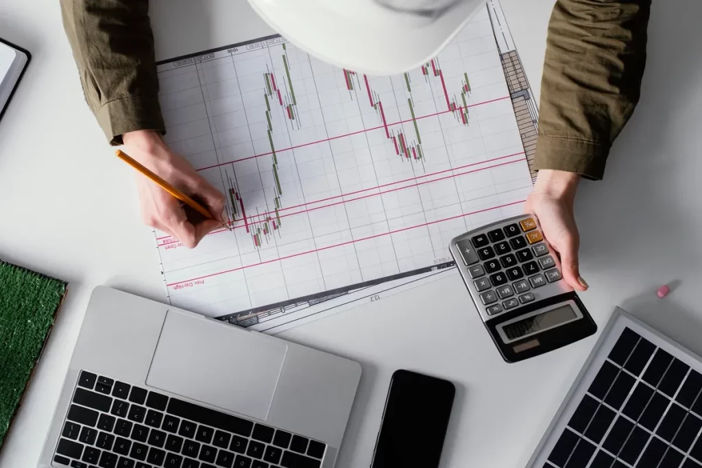Pessoa analisando gráficos financeiros impressos, segurando uma calculadora e fazendo anotações, com laptop e celular ao redor.