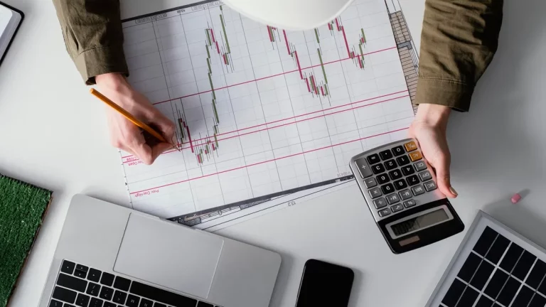 Pessoa analisando gráficos financeiros impressos, segurando uma calculadora e fazendo anotações, com laptop e celular ao redor.