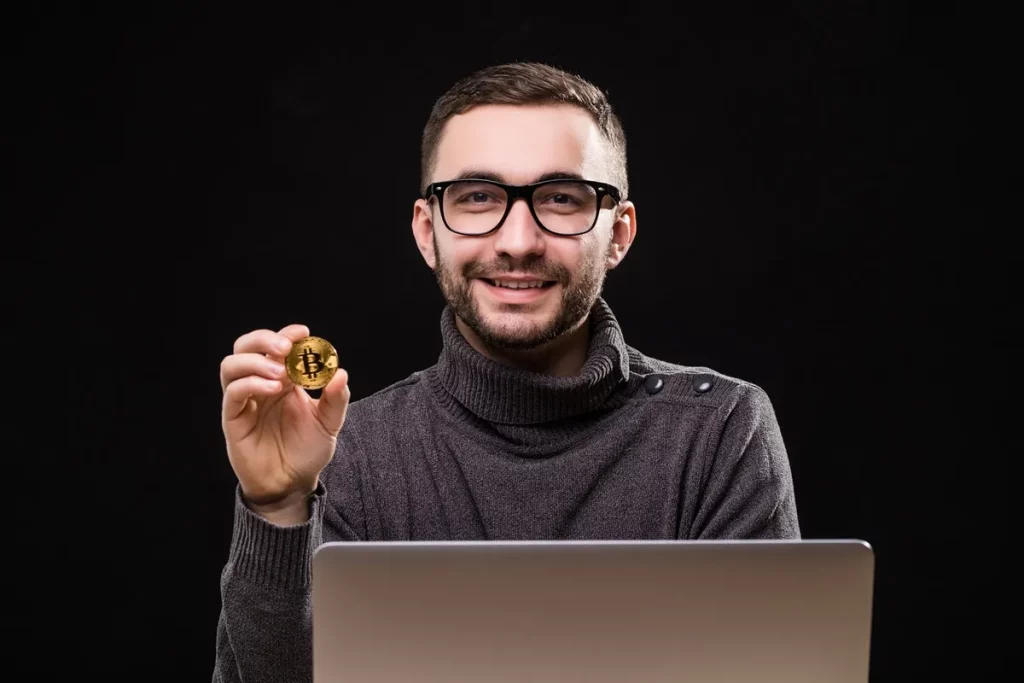 Homem sorridente com óculos e suéter cinza segurando uma moeda de Bitcoin em frente a um notebook com fundo preto.