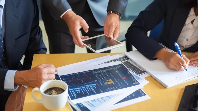 Executivos analisando gráficos financeiros e dados de mercado em um escritório, com documentos impressos e um tablet, representando tomada de decisões estratégicas em investimentos.