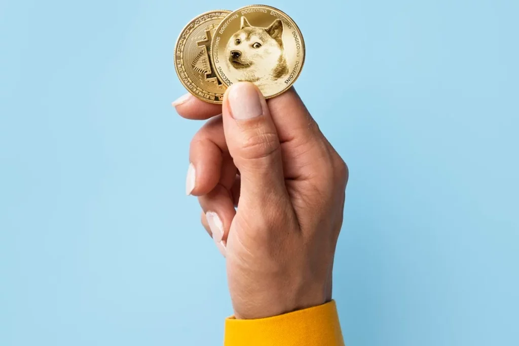 Pessoa segurando duas moedas físicas representando criptomoedas, incluindo a Dogecoin, com fundo azul claro.