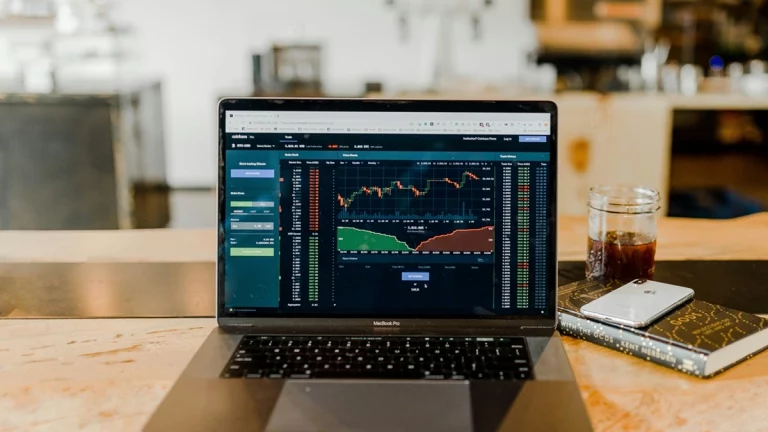 Laptop exibindo plataforma de trading com gráficos de mercado financeiro, ao lado de um café e um smartphone sobre a mesa.