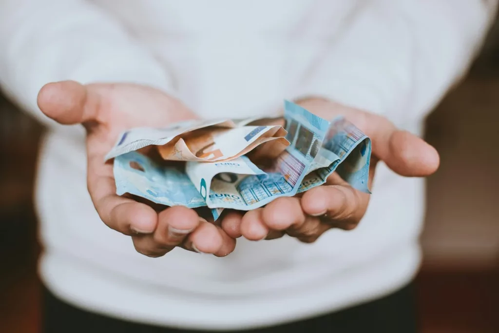Mãos abertas segurando várias cédulas de euro amassadas, sugerindo decisão financeira ou ato de dar/receber dinheiro.