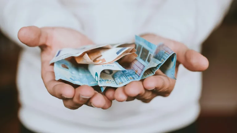 Mãos abertas segurando várias cédulas de euro amassadas, sugerindo decisão financeira ou ato de dar/receber dinheiro.