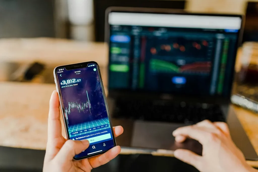 Pessoa segurando smartphone com gráfico do Bitcoin e valor de cotação, enquanto opera plataforma de investimentos em notebook ao fundo.