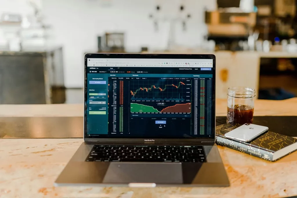 Notebook com tela exibindo plataforma de trading com gráficos e ordens de compra e venda, sobre mesa com livro, celular e copo com bebida