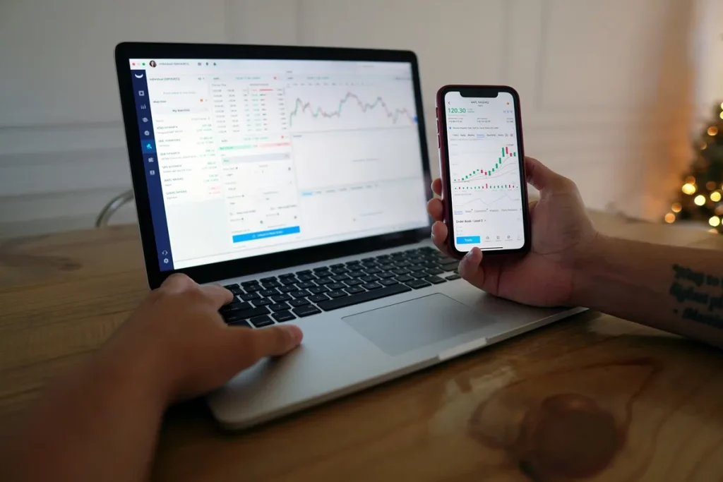 Pessoa analisando gráficos de ações em um notebook e smartphone simultaneamente, com interface de plataforma de investimentos