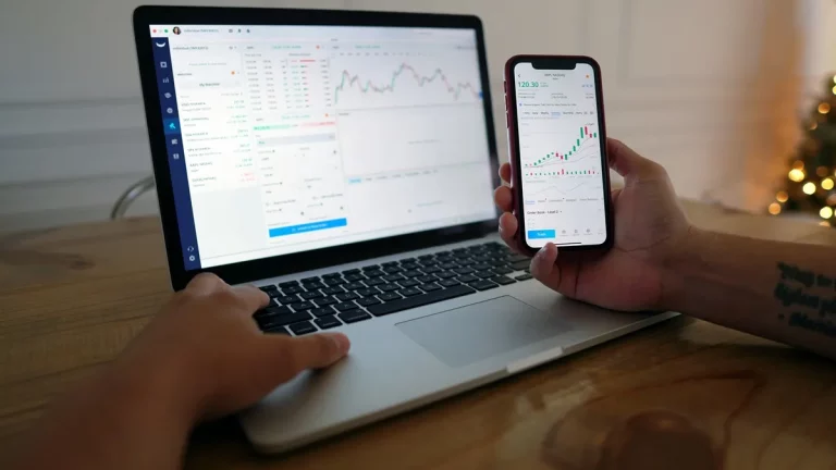 Pessoa analisando gráficos de ações em um notebook e smartphone simultaneamente, com interface de plataforma de investimentos