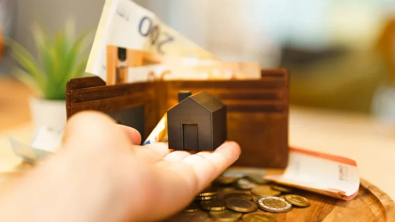 Mão segurando uma miniatura de casa em frente a uma carteira aberta com cédulas de euro e moedas sobre a mesa, simbolizando planejamento financeiro para morar sozinho.