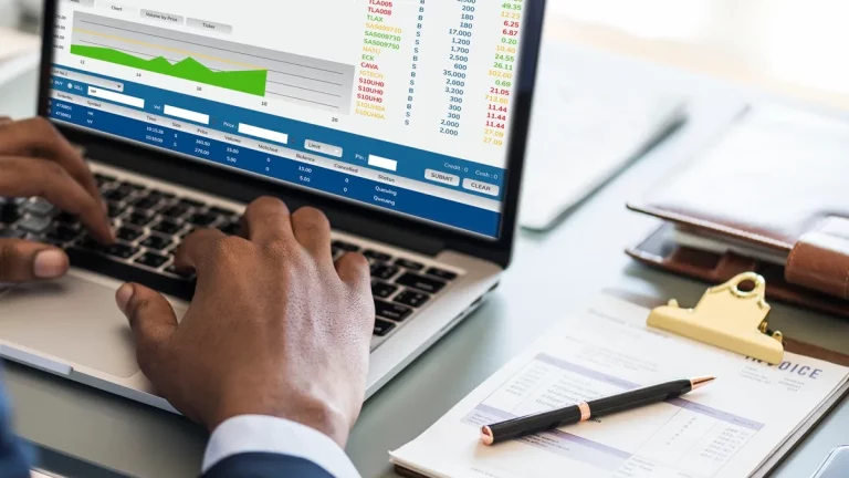 Homem analisando gráficos e cotações na tela do notebook enquanto realiza operações financeiras, com nota fiscal e caneta sobre a mesa.