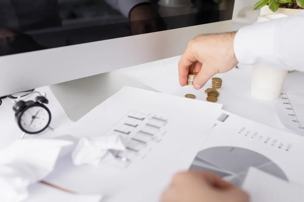 Imagem de uma mão organizando pilhas de moedas sobre papéis financeiros e gráficos, com um relógio de mesa e uma planta ao fundo. O computador está parcialmente visível, indicando um ambiente de trabalho financeiro.