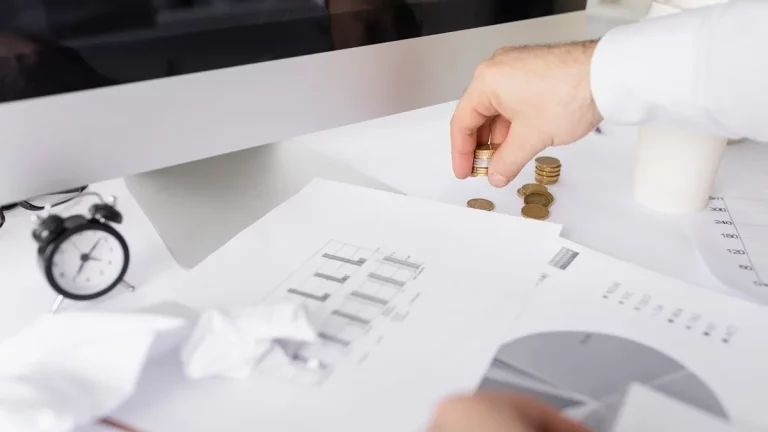 Imagem de uma mão organizando pilhas de moedas sobre papéis financeiros e gráficos, com um relógio de mesa e uma planta ao fundo. O computador está parcialmente visível, indicando um ambiente de trabalho financeiro.