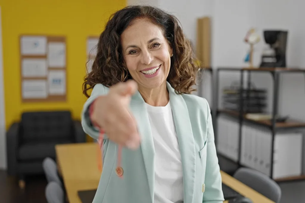 Mulher madura sorridente estendendo a mão em gesto de cumprimento, vestindo blazer verde-claro em escritório com fundo amarelo