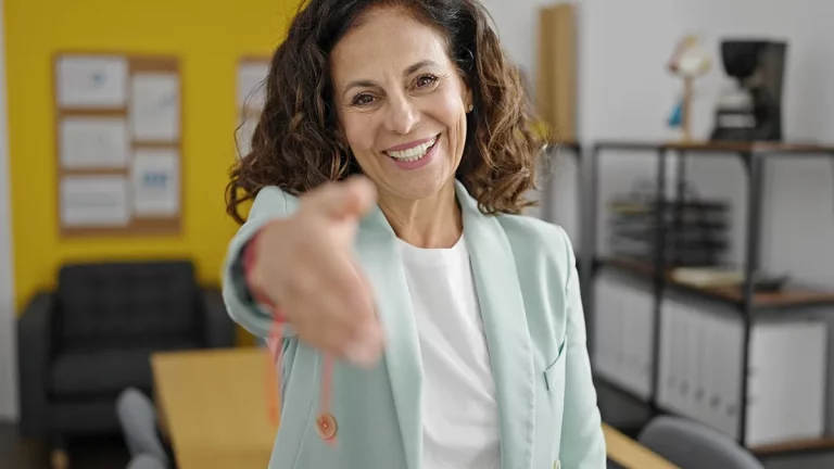 Mulher madura sorridente estendendo a mão em gesto de cumprimento, vestindo blazer verde-claro em escritório com fundo amarelo