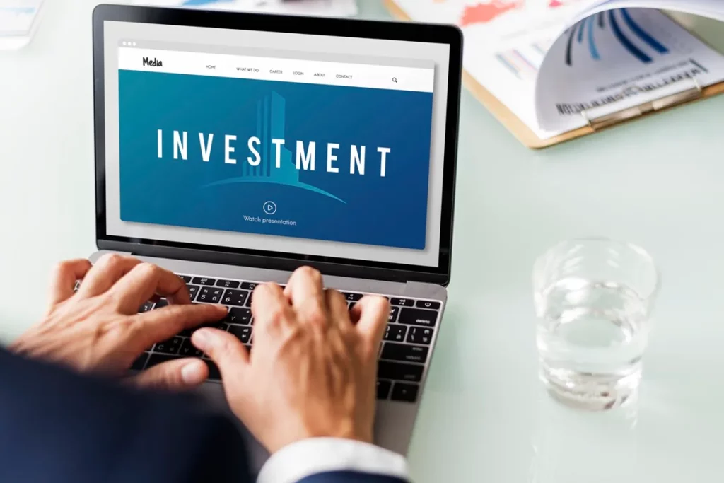 Pessoa digitando em laptop com tela exibindo a palavra "INVESTMENT" e documentos financeiros ao fundo.