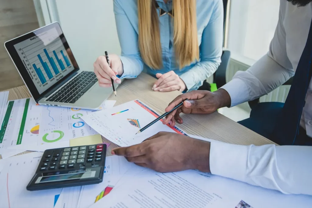 Imagem de duas pessoas analisando gráficos e relatórios financeiros, com um laptop aberto mostrando gráficos de barras. Uma pessoa está usando um lápis para fazer anotações em papéis enquanto a outra aponta para os gráficos.