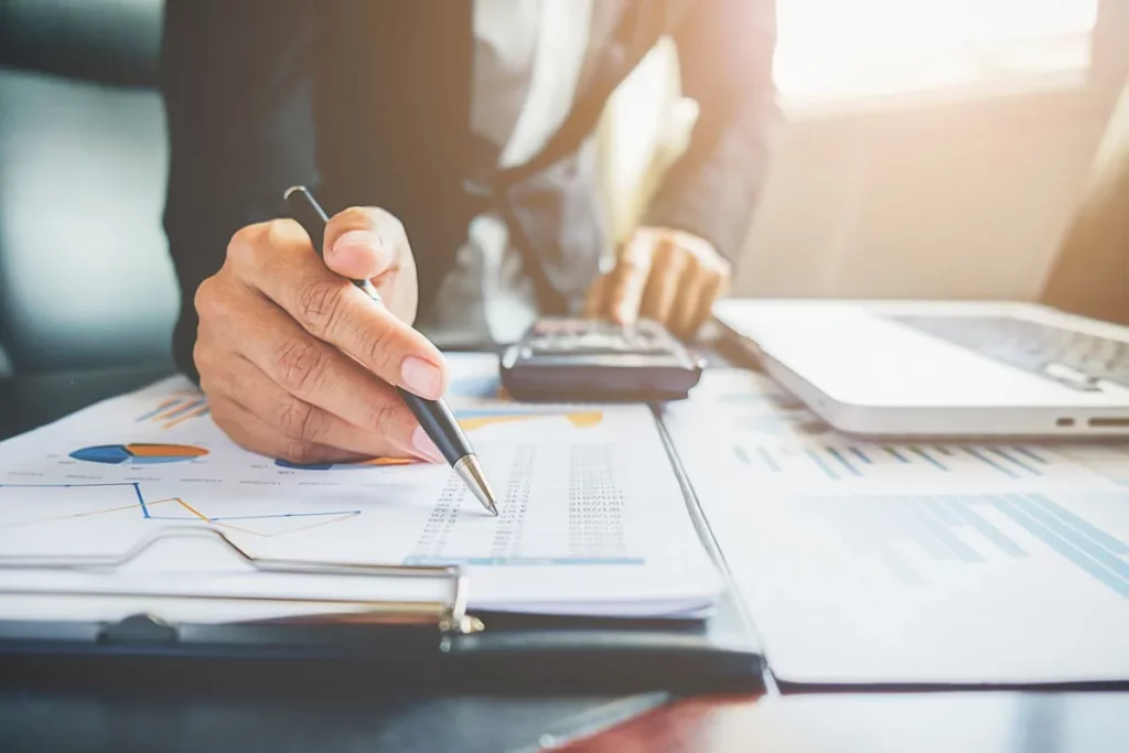 Executivo revisando relatórios financeiros, gráficos e planilhas em uma mesa de escritório com calculadora e laptop.