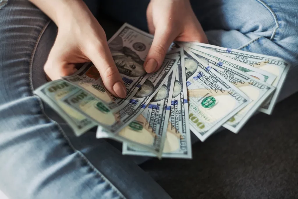 Mãos segurando diversas notas de 100 dólares em formato de leque, simbolizando riqueza, lucro e organização financeira.