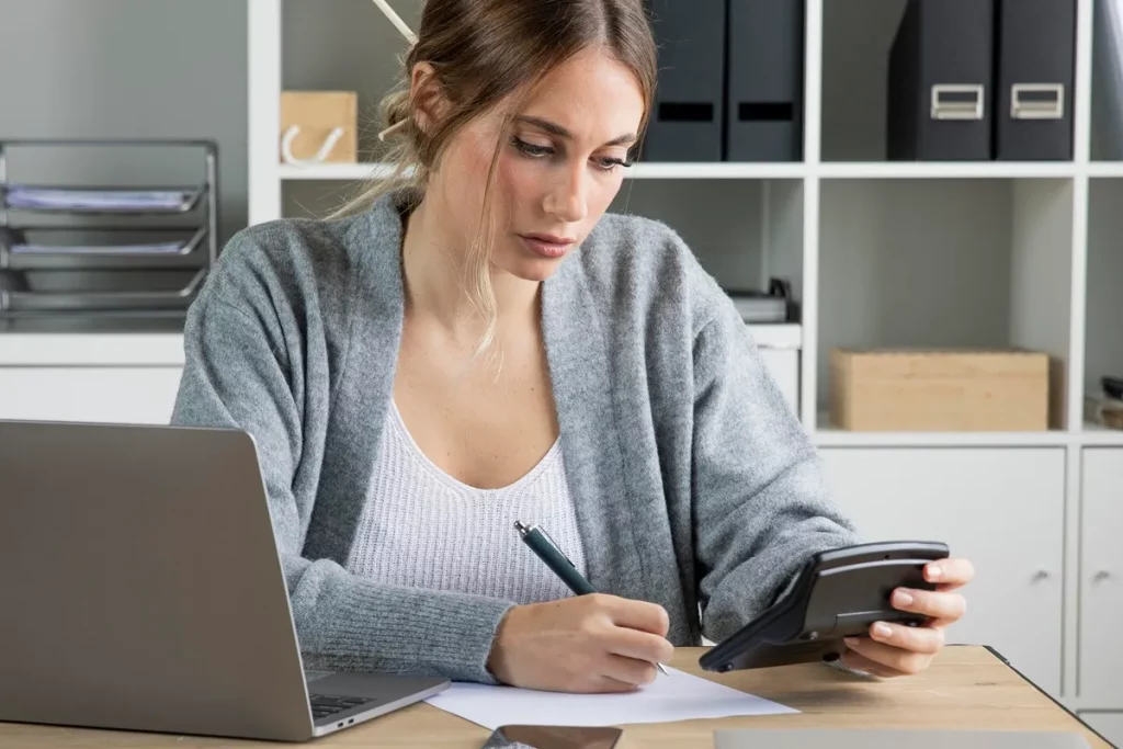 Imagem de uma mulher usando calculadora e caneta, preenchendo um formulário DARF sobre a mesa de trabalho, com laptop ao fundo