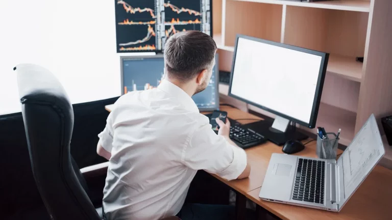 Homem de costas sentado em cadeira de escritório, analisando gráficos financeiros em três telas de computador e segurando um smartphone ao lado de um notebook aberto